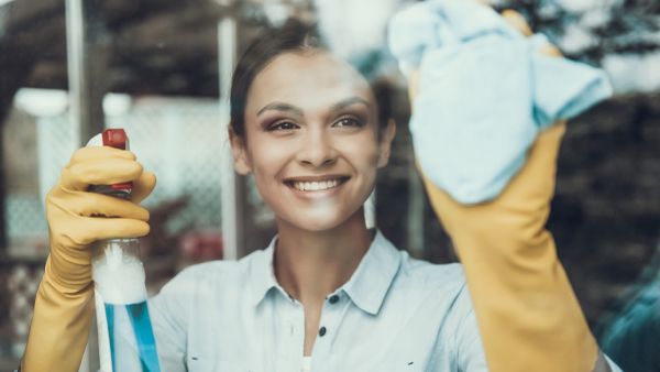 Smiling woman wearing yellow gloves cleaning a window with spray and cloth during spring cleaning.
