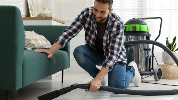 Man vacuuming under a sofa with professional cleaning equipment as part of spring cleaning and home maintenance.