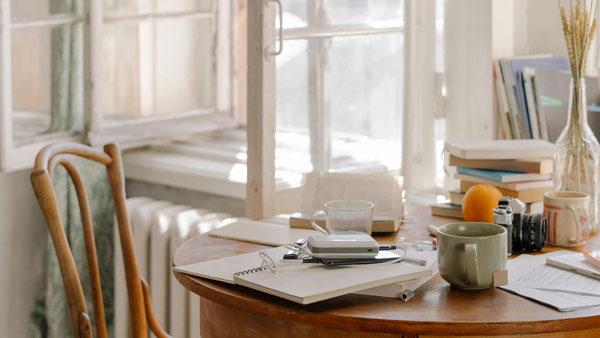 Bright cozy home workspace with books, notebooks, and coffee mugs on a wooden table beside an open window letting in fresh air.